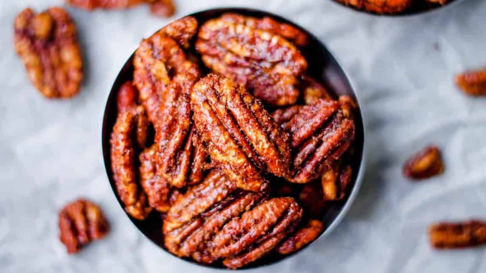 A bowl filled with candied pecans on a light surface, with a few pecans scattered around the bowl. The nuts are glossy and appear to be coated in a sugar glaze.