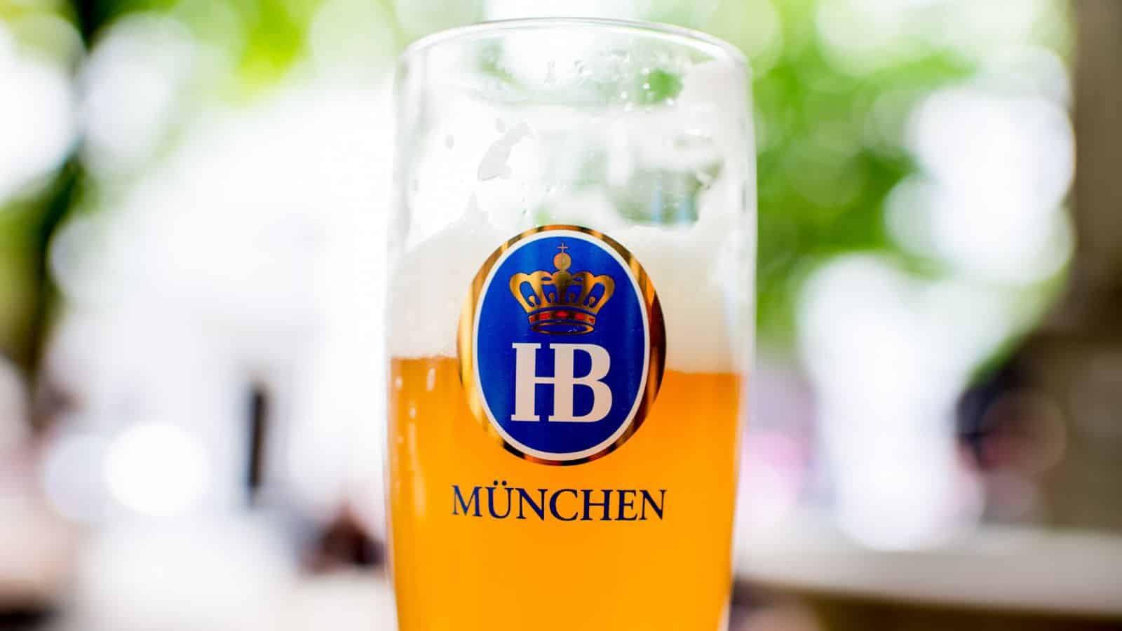 A tall glass of beer with a frothy head, featuring the HB M&uuml;nchen logo on the front. The background is blurred greenery, suggesting an outdoor setting.
