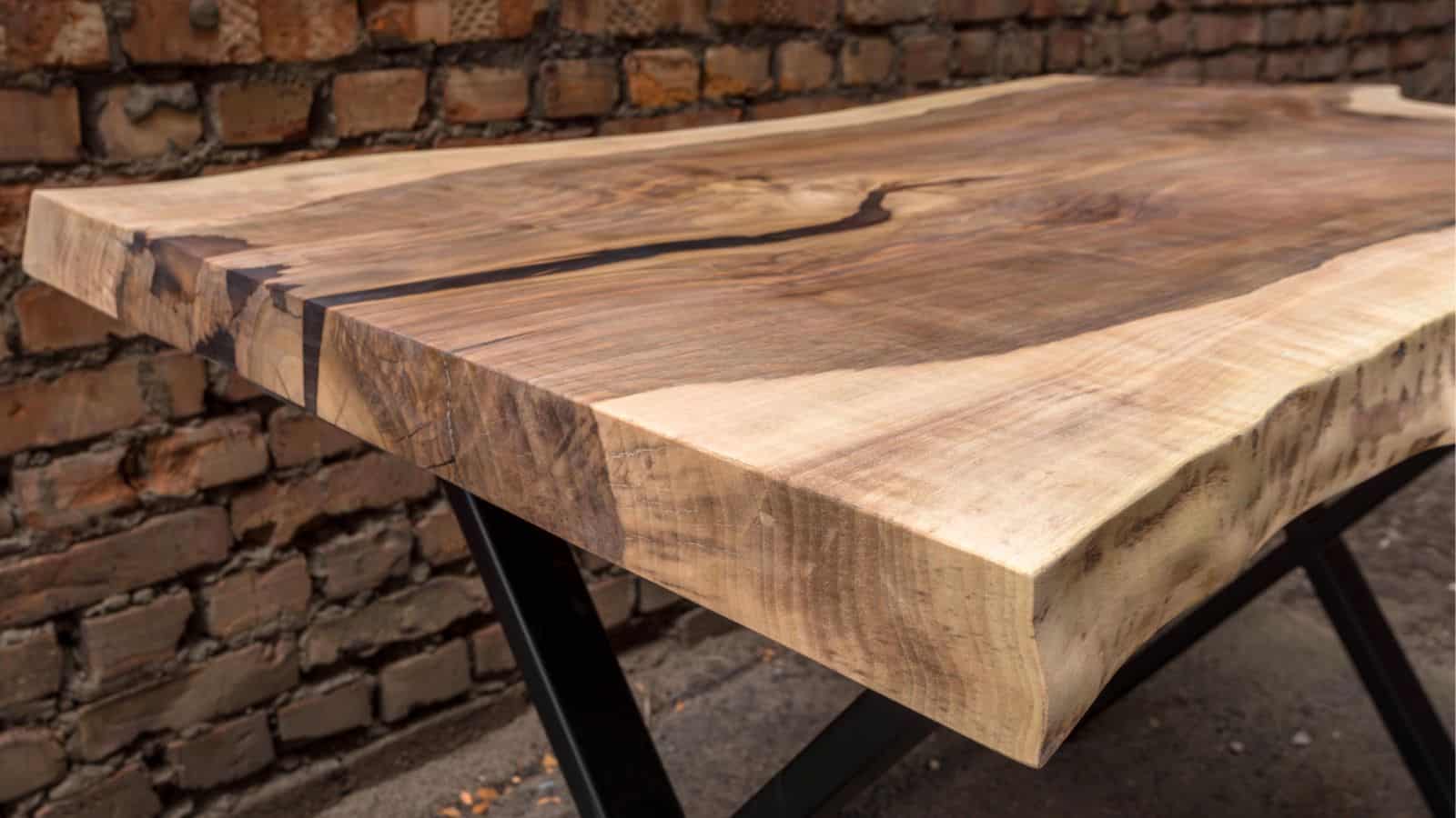 A close-up of a thick wooden table with a natural edge, displaying its smooth grain and unique texture. The table is supported by metal legs. In the background, there is a brick wall.
