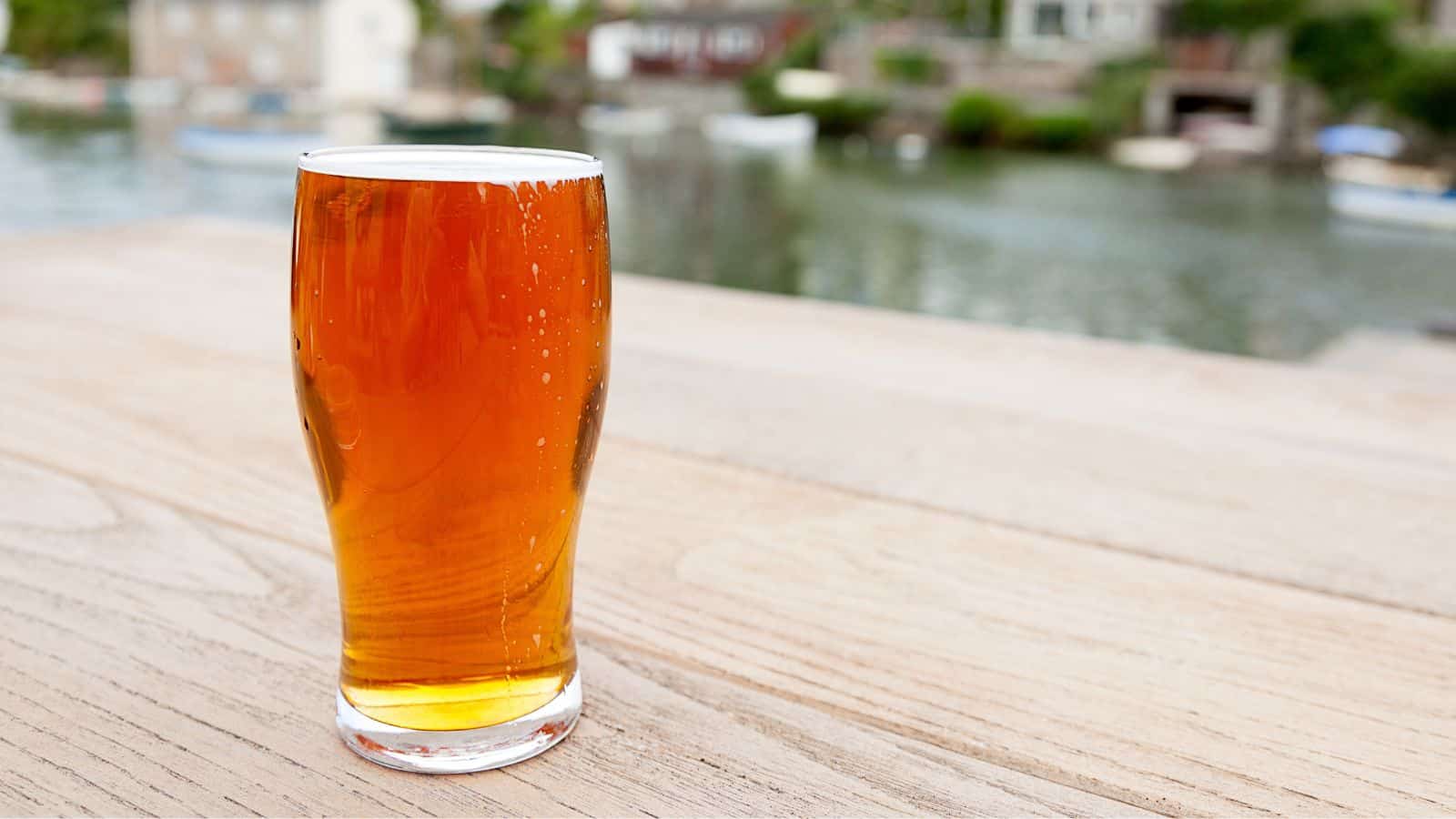 A pint of amber-colored beer in a clear glass sits on a wooden table outdoors. The background features a blurred view of water with small boats and houses, suggesting a waterfront setting.
