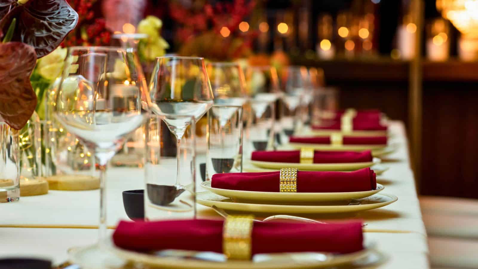 A elegant dining table setup with neatly arranged wine glasses, water glasses, and plates. Each place setting features a red napkin with a gold napkin ring. The background is softly lit with warm lights and floral decorations.