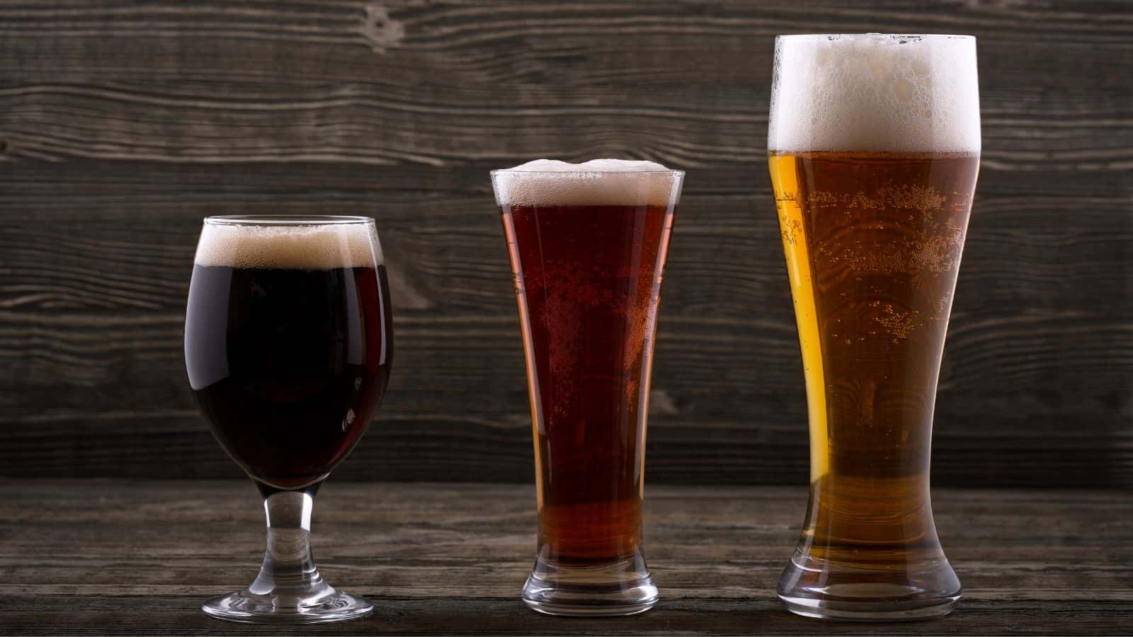 Three glasses of beer on a wooden surface against a dark wooden background. The beers vary in color and size, with the left glass holding dark beer, the middle glass a reddish-brown beer, and the right glass a light golden beer.