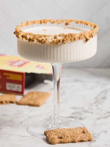 A creamy cocktail in a ribbed glass with a crumb-coated rim sits on a marble surface. Nearby are a cinnamon stick, speculoos cookies, a red box, and two liquor bottles partially visible in the background.