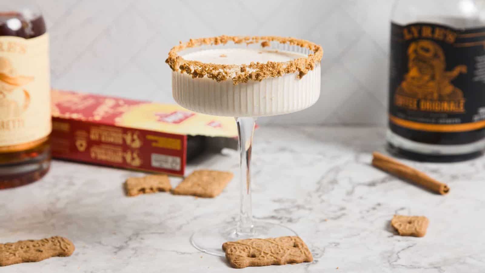 A creamy cocktail in a ribbed glass with a crumb-coated rim sits on a marble surface. Nearby are a cinnamon stick, speculoos cookies, a red box, and two liquor bottles partially visible in the background.