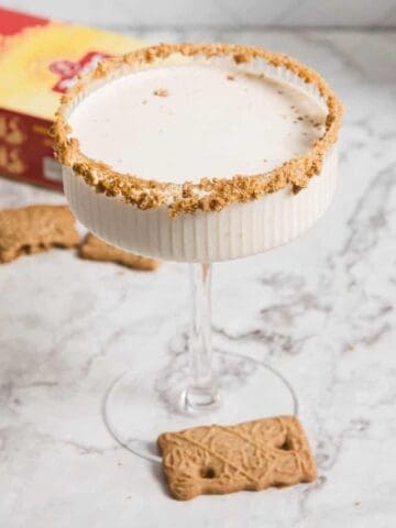 A glass with a beige creamy drink sits on a marble surface. The glass has a crushed cookie rim. Surrounding the glass are several speculoos cookies, a cinnamon stick, and two bottles of liqueur. A red book is in the background.