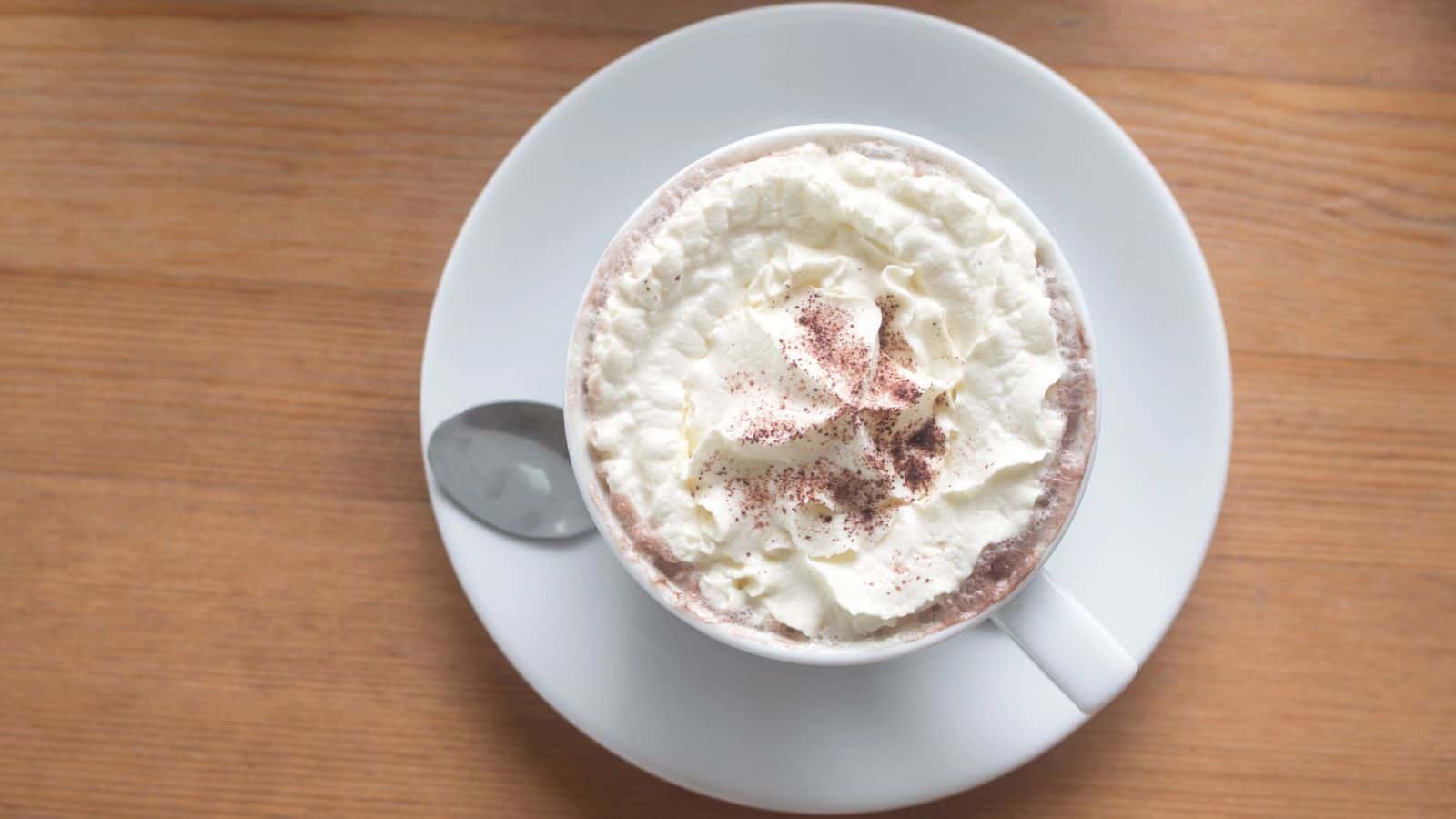 A white cup filled with hot chocolate topped with whipped cream and sprinkled with cocoa powder, resting on a white saucer with a silver spoon, placed on a wooden table.
