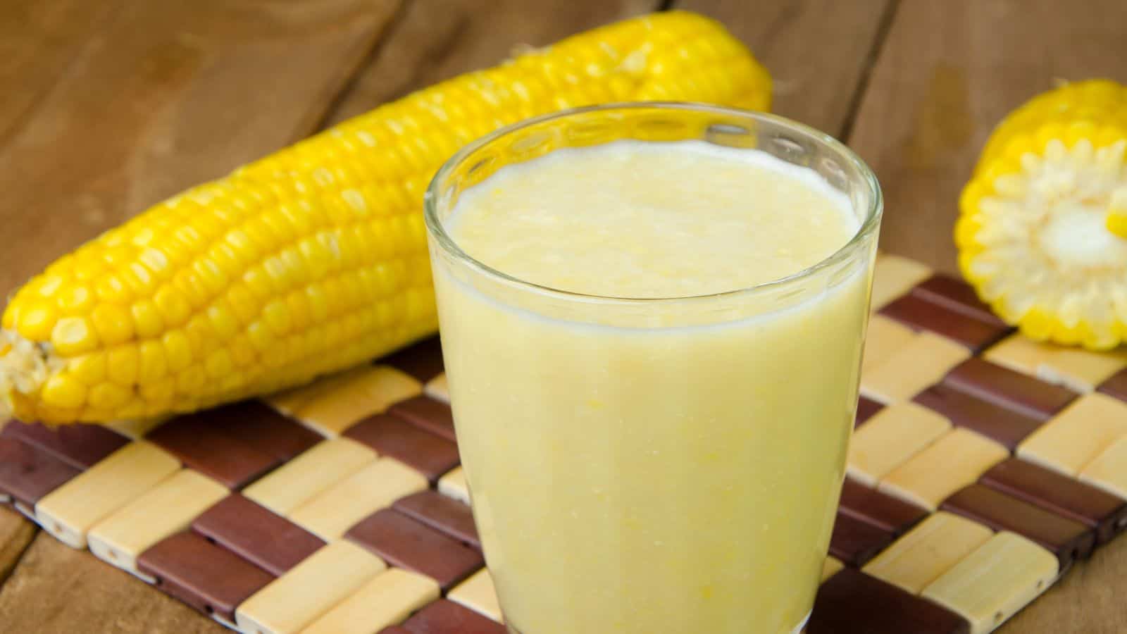 A glass of corn juice is placed on a woven mat, with two ears of corn, one whole and one partially visible, in the background. The wooden surface beneath the mat adds a rustic touch.