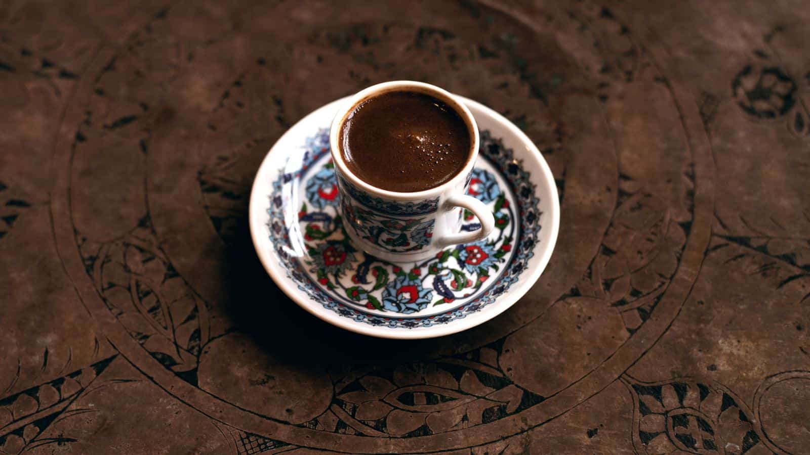 A small, ornately decorated cup filled with dark coffee sits on a matching saucer. The cup and saucer feature intricate floral patterns in blue, red, and green. They are placed on a textured, patterned surface.