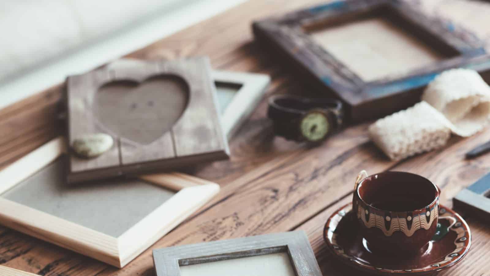 A wooden table holds several empty picture frames, a patterned teacup with a saucer, a vintage-style watch, and a decorative rock shaped like a heart. Rolled-up paper is also visible in the background.
