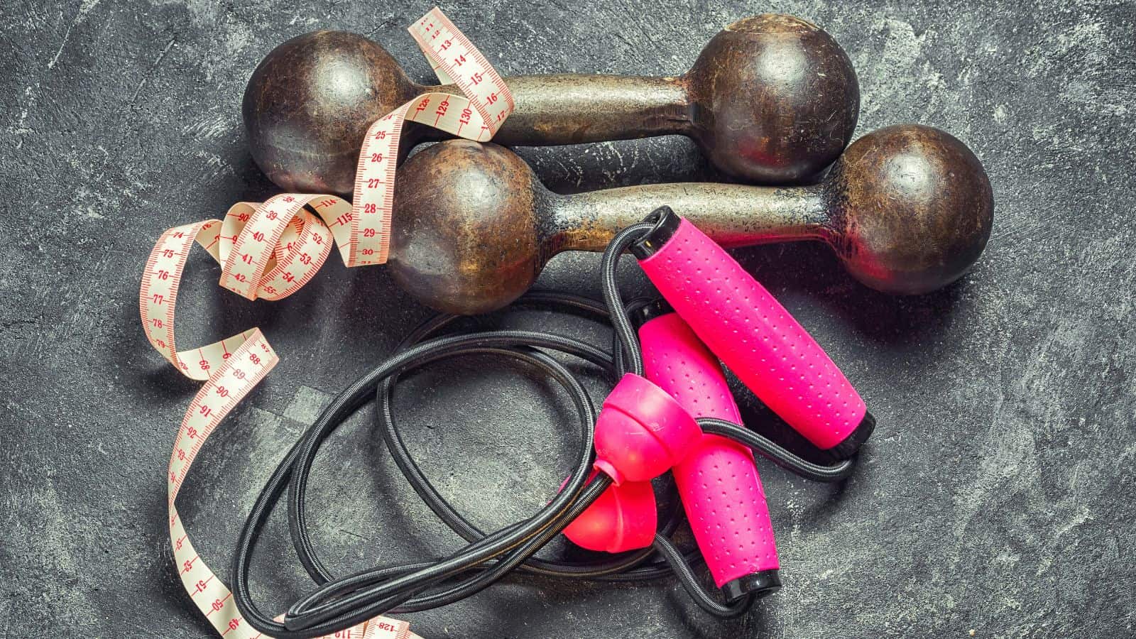 A pair of rustic dumbbells, a measuring tape, and a pink jump rope with black handles are arranged on a dark, textured surface.