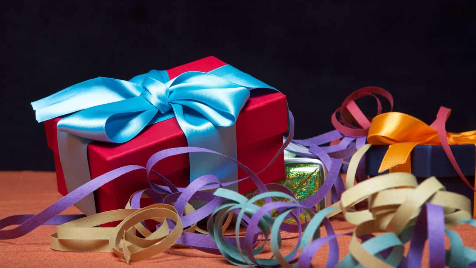 A red gift box with a blue ribbon is surrounded by colorful streamers. A smaller box with an orange ribbon is partially visible in the background, sitting on a textured orange surface against a dark backdrop.