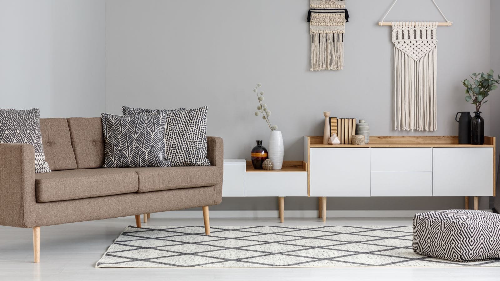 A modern living room featuring a brown sofa with patterned cushions, a white and wood console with decorative items, and macram&eacute; wall hangings. A geometric rug and a matching pouf sit on the light-colored floor.