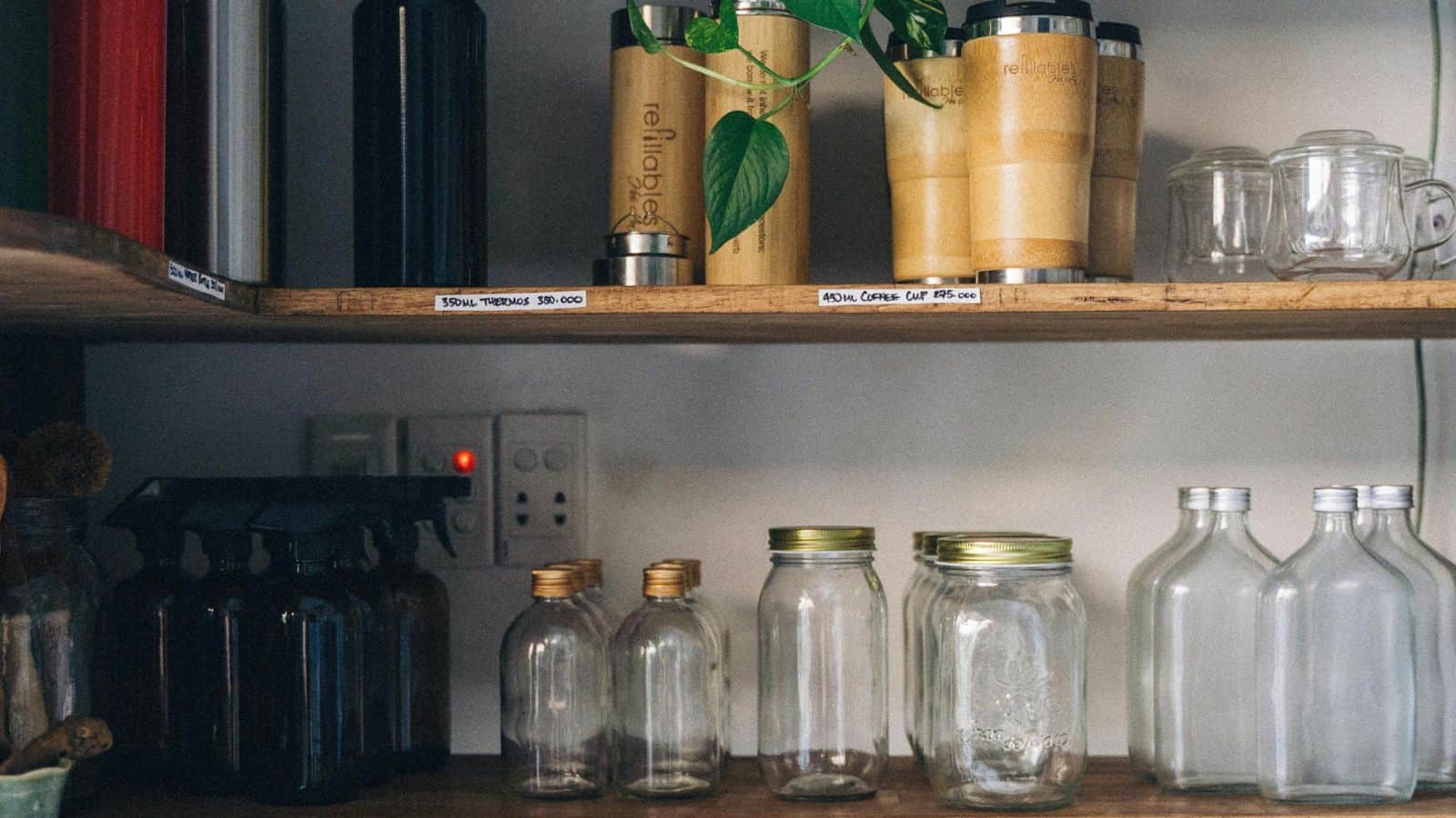 A wooden shelf holds reusable containers, including bamboo and metal travel mugs on the upper shelf, and glass jars and bottles on the lower shelf. A plant hangs from above, and there are labels and a light switch nearby.