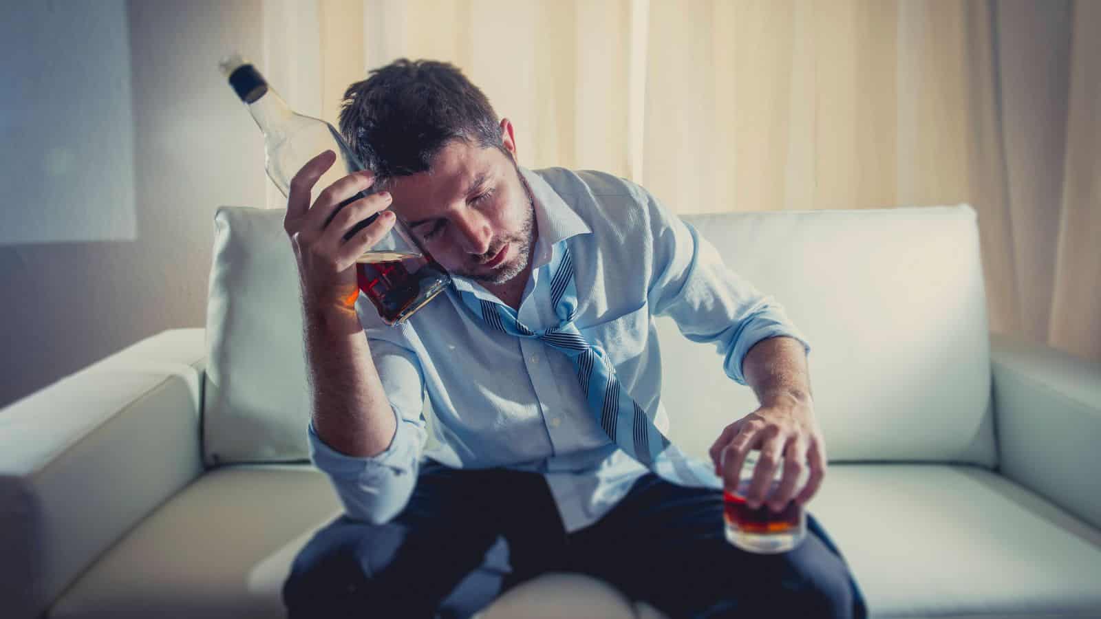 A man with disheveled hair and wearing a blue shirt and tie sits on a white couch holding a bottle and a glass of dark liquid. His eyes are closed and his head rests against the bottle. The background features light-colored curtains.