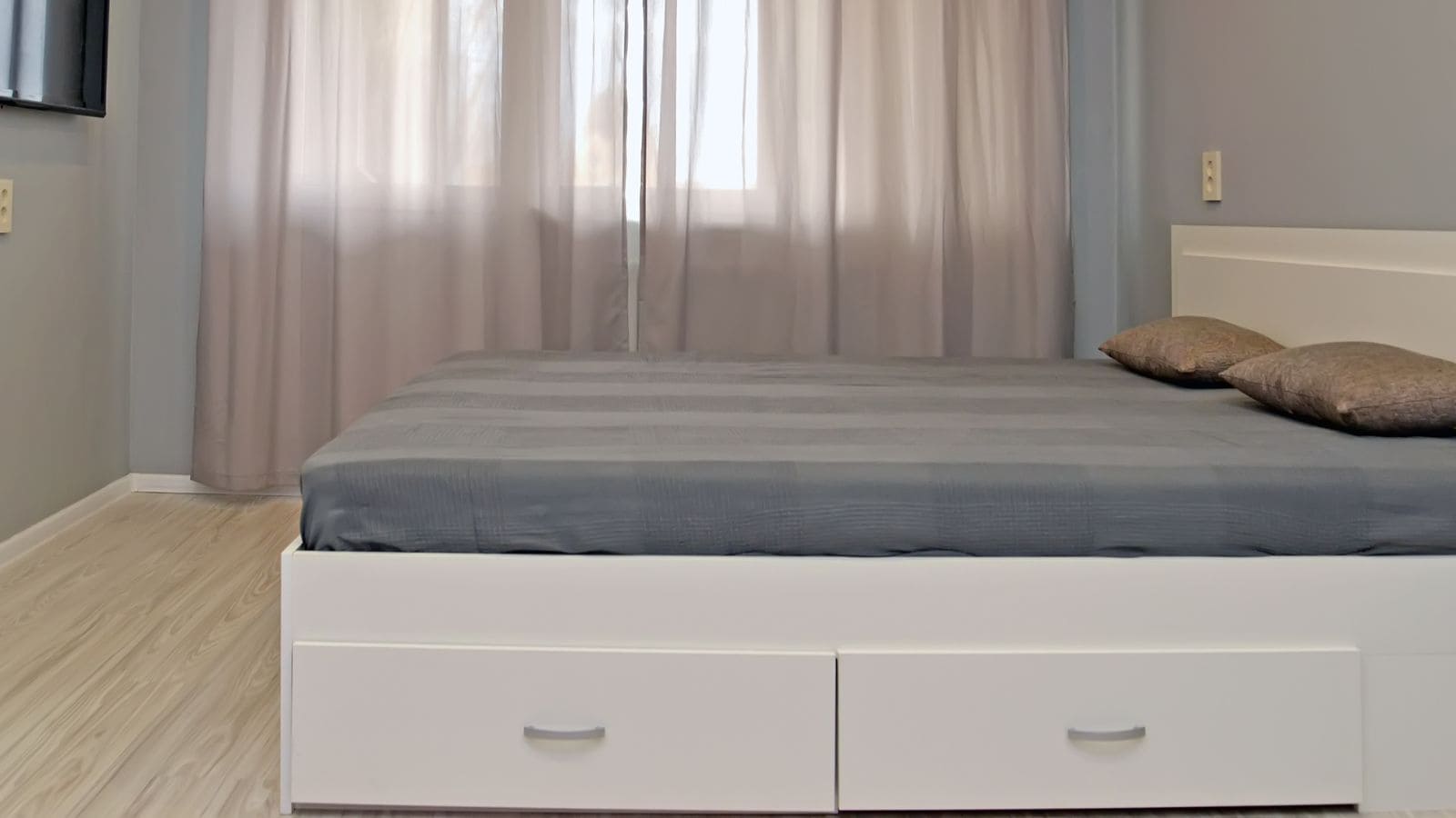 A minimalist bedroom with a large bed featuring a grey cover and two brown pillows. The bed has a white base with drawers. Sheer curtains cover the window, and the walls are painted in a neutral gray tone. Light wooden flooring is visible.