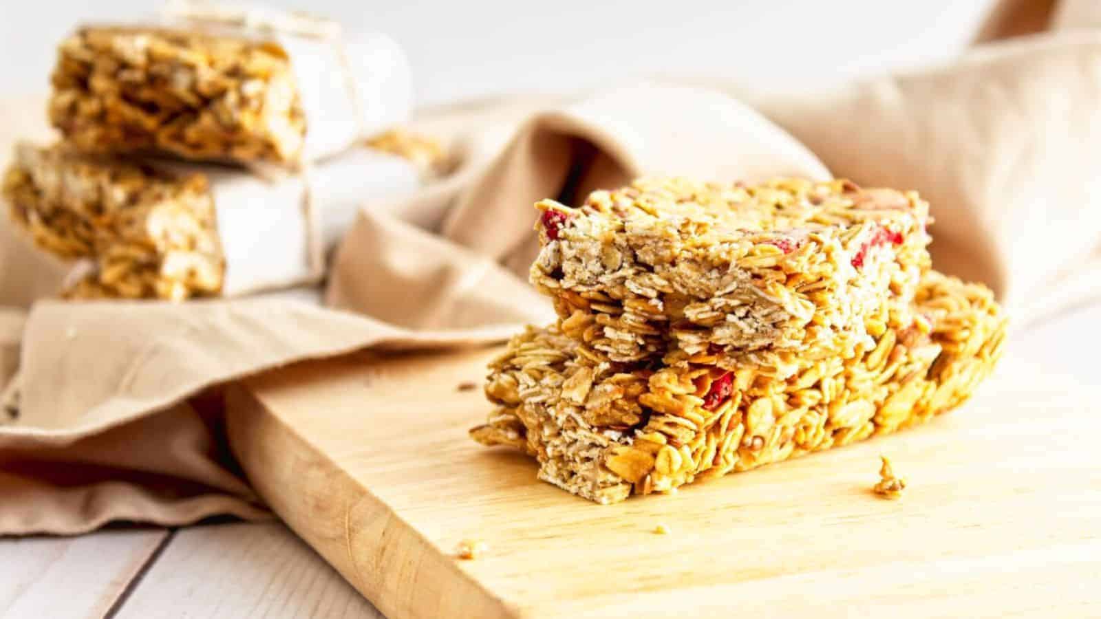 A close-up of two granola bars stacked on a wooden board. The bars are made of oats and appear to contain dried fruit. In the background, additional wrapped granola bars are slightly out of focus, with a beige cloth underneath.