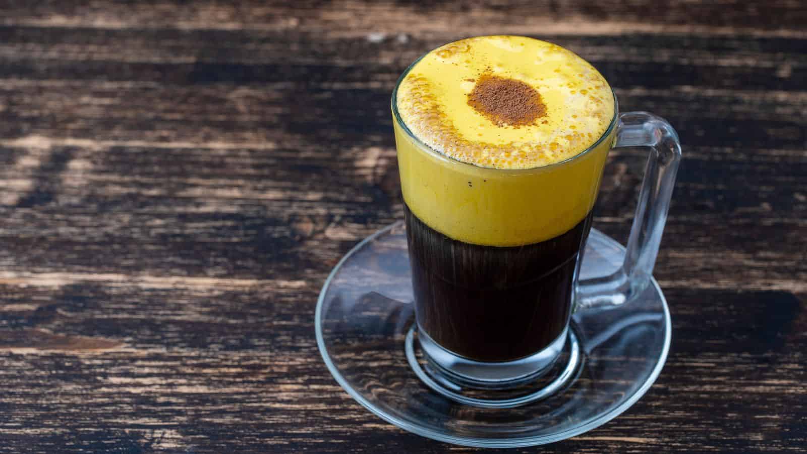 A glass mug of black coffee topped with a thick layer of yellow, frothy egg cream, sprinkled with cinnamon, sits on a glass saucer. The mug is placed on a wooden table.