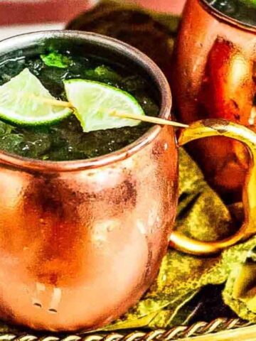 Two copper mugs filled with a dark beverage, garnished with lime wedges on skewers, rest on a decorative brass tray with a fabric lining. The mugs have a frosty exterior, suggesting the drink is cold.