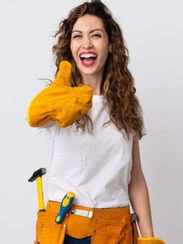 A woman wearing a white T-shirt and a tool belt gives a thumbs-up gesture. She is smiling and wearing yellow work gloves. There are tools like a hammer and a screwdriver in her belt. The background is plain white.