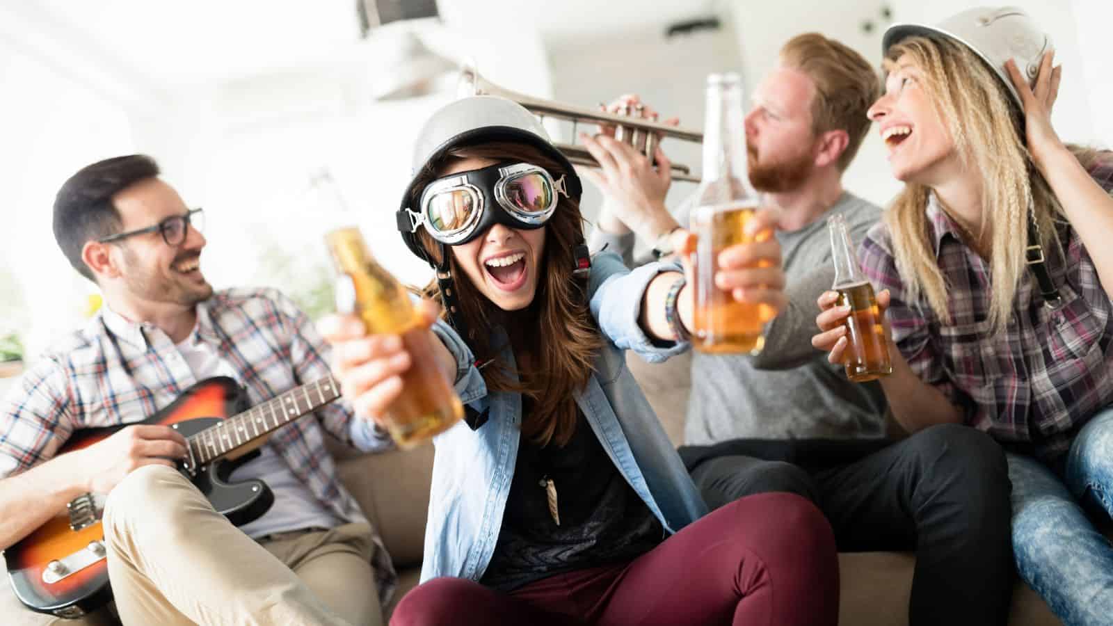 A group of four people are having fun indoors. One person is holding bottles and wearing goggles, another is playing a guitar, a third is wearing a helmet and holding a beer, and the fourth is playing a trumpet. They appear to be enjoying a lively moment.