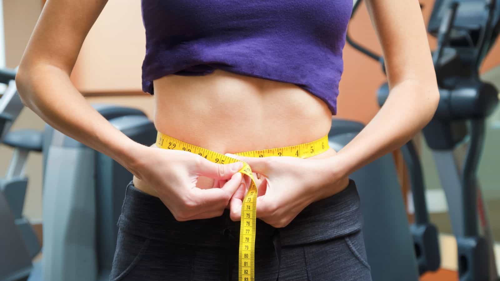 A person in a purple crop top and black pants measures their waist with a yellow measuring tape in a gym setting, with exercise equipment visible in the background.