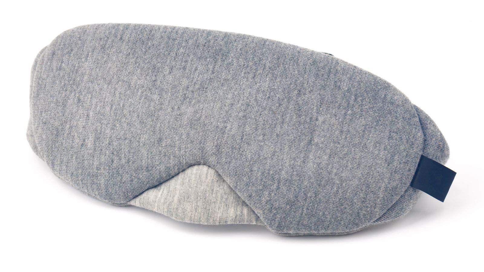 A soft, gray sleep mask is displayed against a white background. The fabric has a textured appearance, and there is a small black tag on one side of the mask.