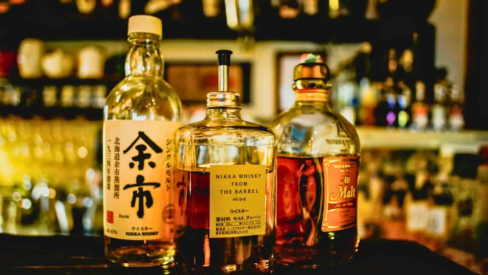 Three bottles of Japanese whisky are displayed on a bar counter. The labels read "Yoichi Single Malt," "Nikka Whisky from the Barrel," and "Taketsuru Pure Malt." Blurred shelves of glasses and additional bottles are visible in the background.