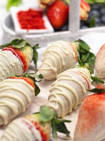 White chocolate-covered strawberries are arranged in rows on a white surface. Some berries are drizzled with more chocolate, while others are dusted with a reddish powder. A container of mixed berries is visible in the background.