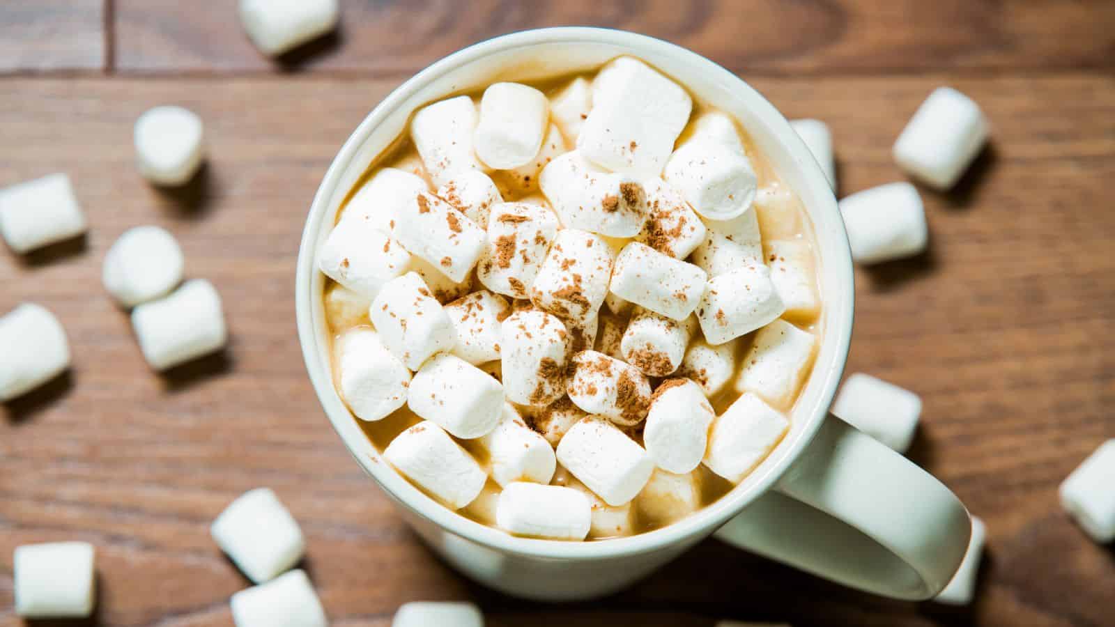 A mug filled with hot chocolate topped with a generous amount of mini marshmallows, sprinkled with cocoa powder. The mug is on a wooden table, surrounded by scattered mini marshmallows.