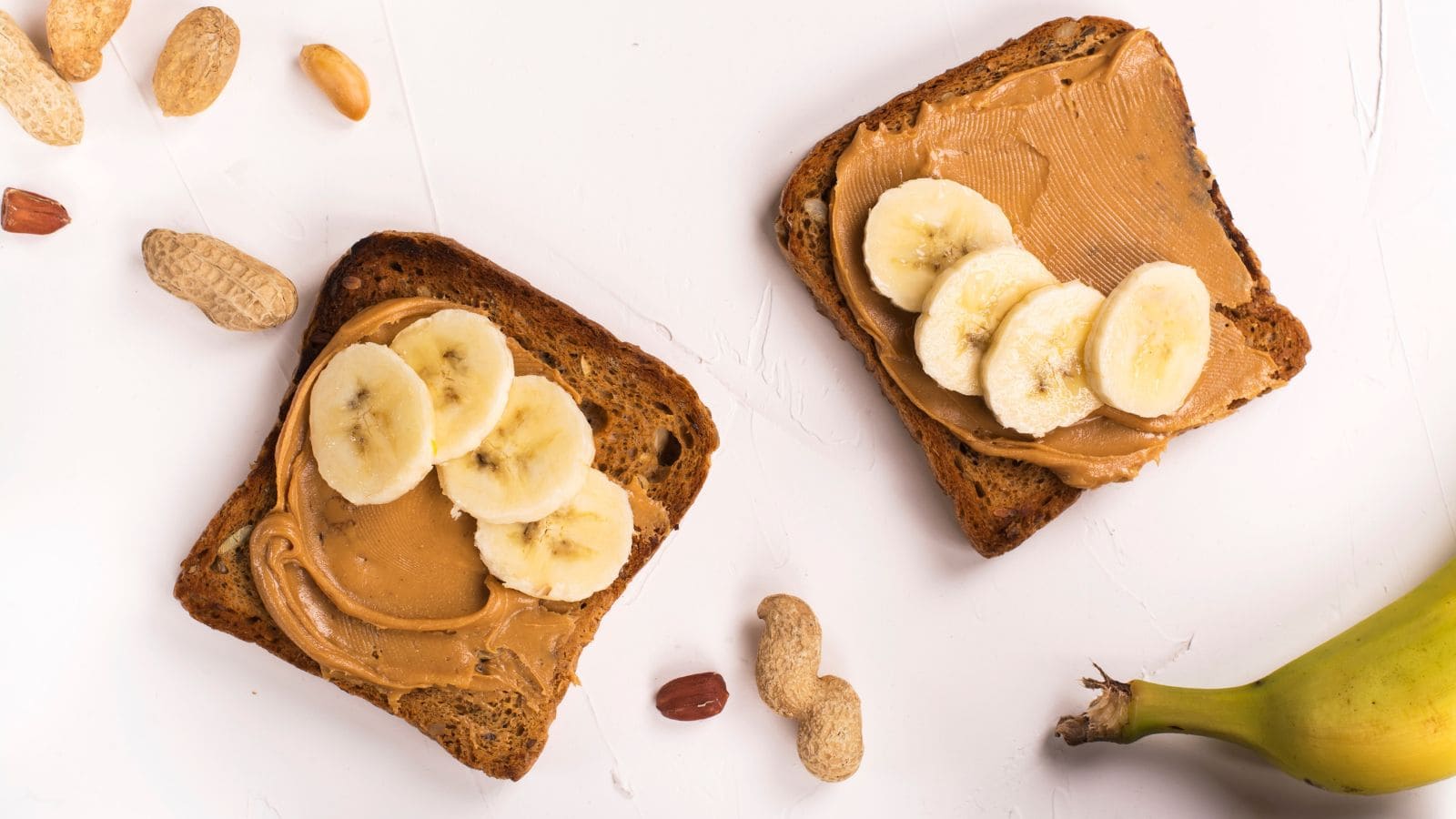 Two slices of toast with peanut butter and banana slices on a white surface. Surrounding the toast are peanuts, a peeled banana, and a whole banana.