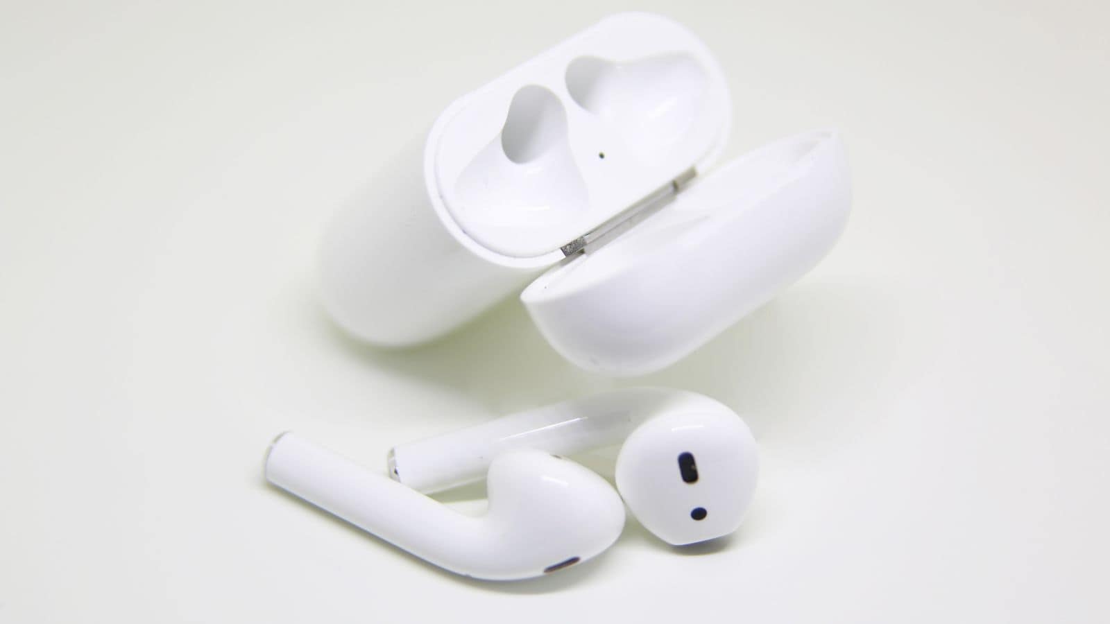 A pair of white wireless earbuds and their open charging case on a white surface. The case is lying flat, and one earbud is resting inside while the other earbud is placed beside it.