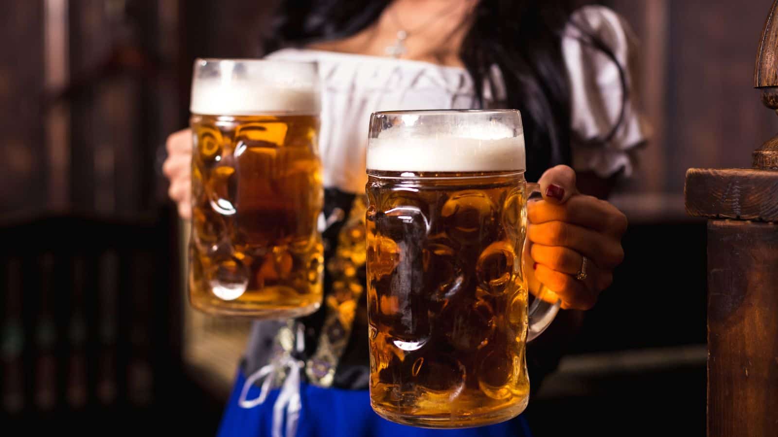 A person in traditional attire holds two large mugs of beer, each topped with foam. The background is a dimly lit wooden interior, possibly a pub or tavern. The focus is on the beer mugs.