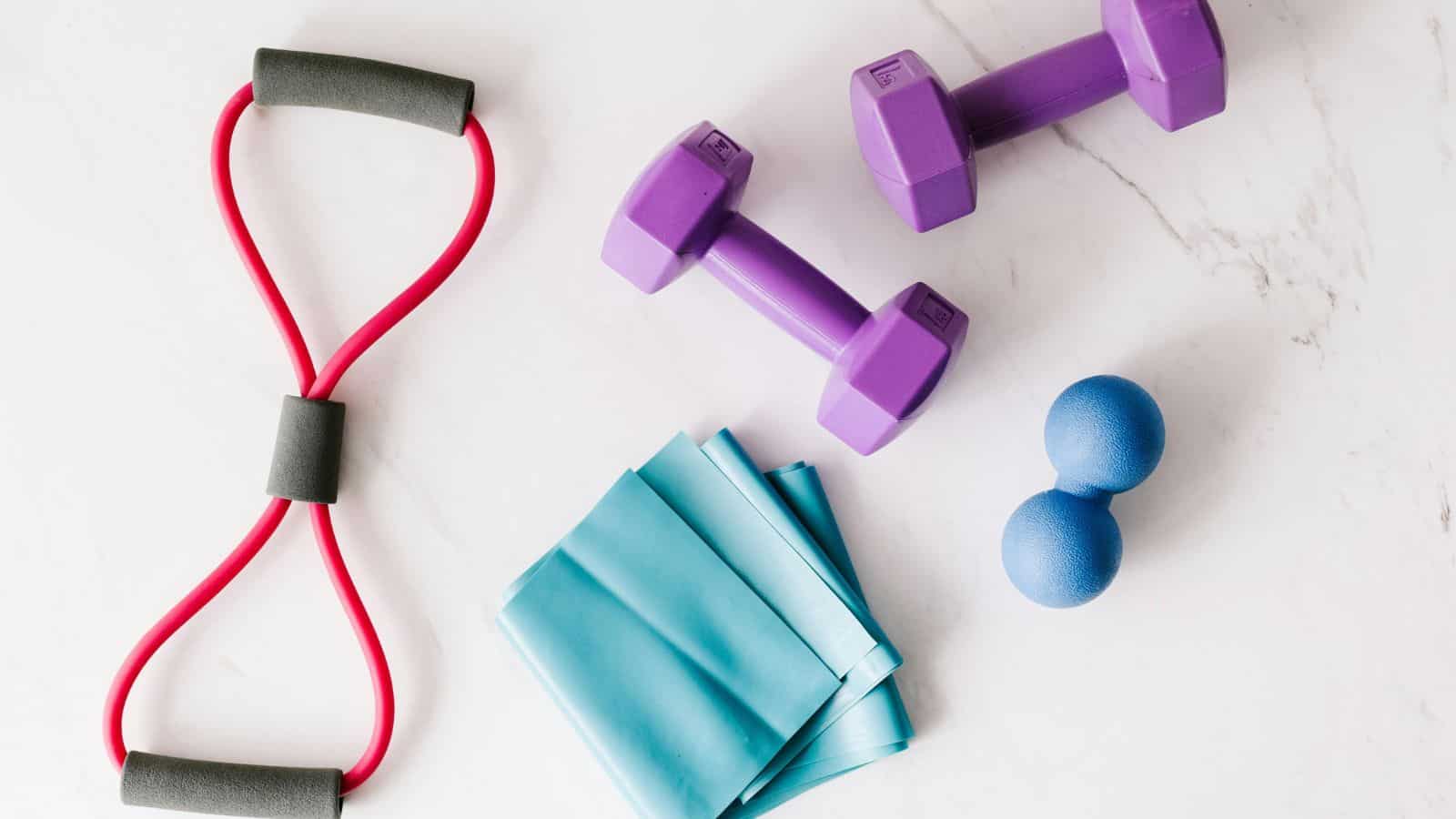 A red resistance band, two purple dumbbells, two blue massage balls, and a folded teal resistance band are arranged on a white marble surface.