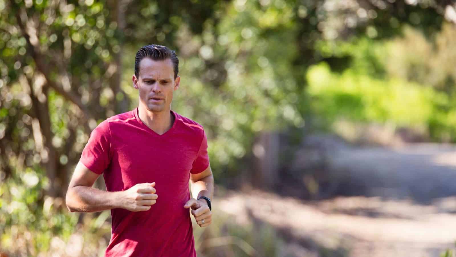 A person in a red shirt is jogging on a dirt path surrounded by green trees and foliage. The scene is outdoors and sunny.