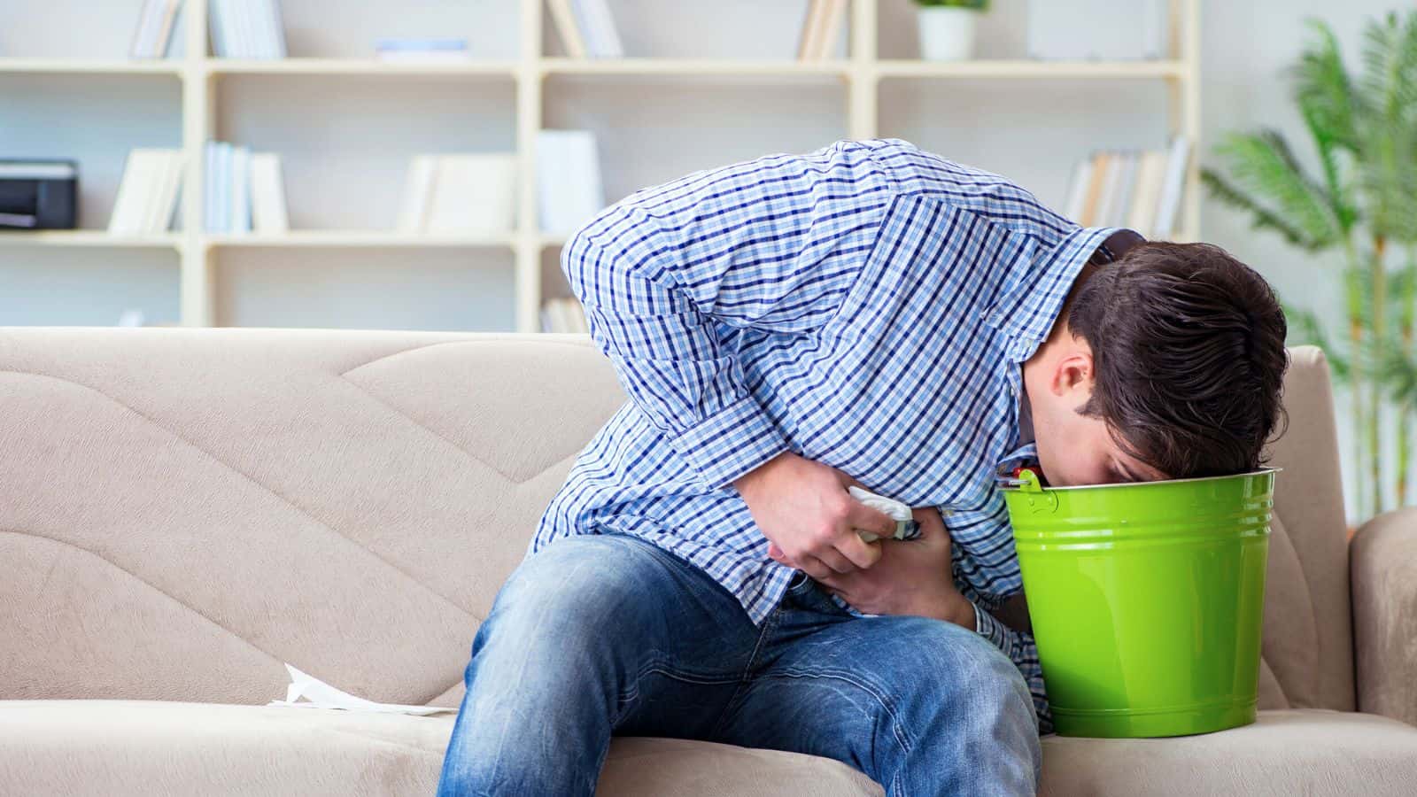 A person in a blue checkered shirt and jeans is sitting on a beige couch, leaning over a green bucket while vomiting. Bookshelves and a potted plant are visible in the background.