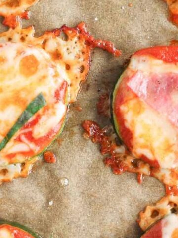 Close-up of baked zucchini slices topped with melted cheese, tomato sauce, and fresh basil on parchment paper. The cheese is slightly browned and bubbly, and the tomato sauce is visible beneath.