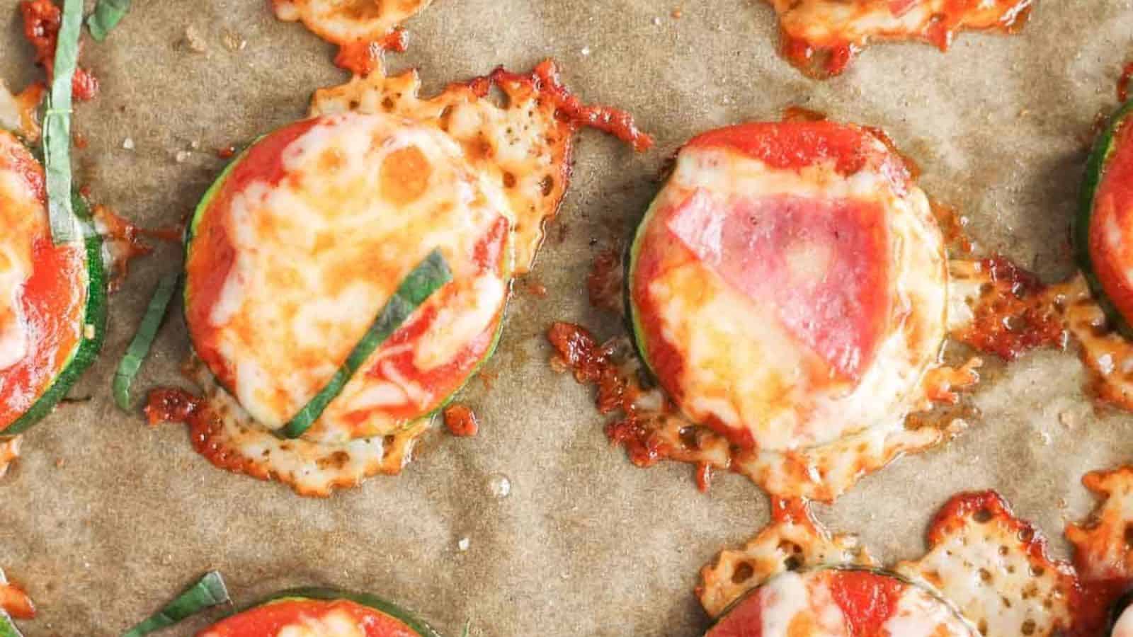 Close-up of baked zucchini slices topped with melted cheese, tomato sauce, and fresh basil on parchment paper. The cheese is slightly browned and bubbly, and the tomato sauce is visible beneath.