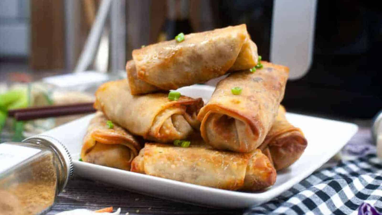 A plate of stacked, golden-brown spring rolls garnished with chopped green onions. A small jar of spices and a checkered cloth are partially visible in the background.
