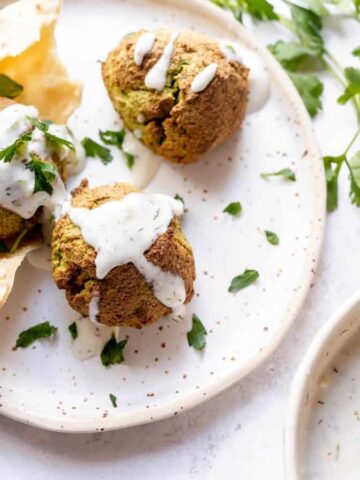 A plate with two falafel balls topped with white sauce and garnished with parsley, accompanied by a piece of pita bread. Beside the plate is a small bowl containing more sauce.