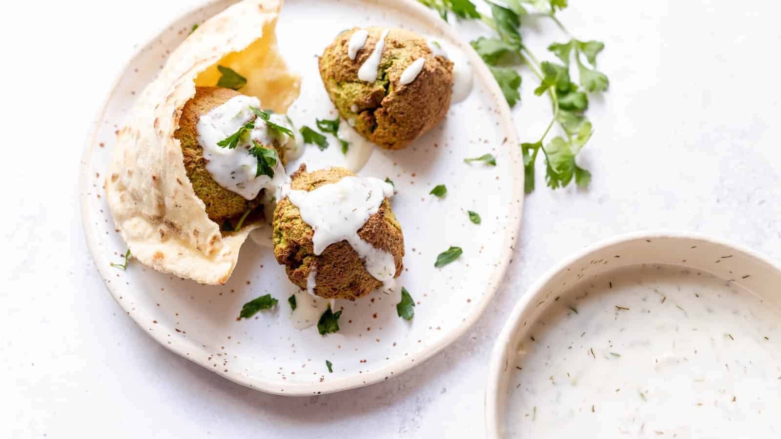 A plate with two falafel balls topped with white sauce and garnished with parsley, accompanied by a piece of pita bread. Beside the plate is a small bowl containing more sauce.