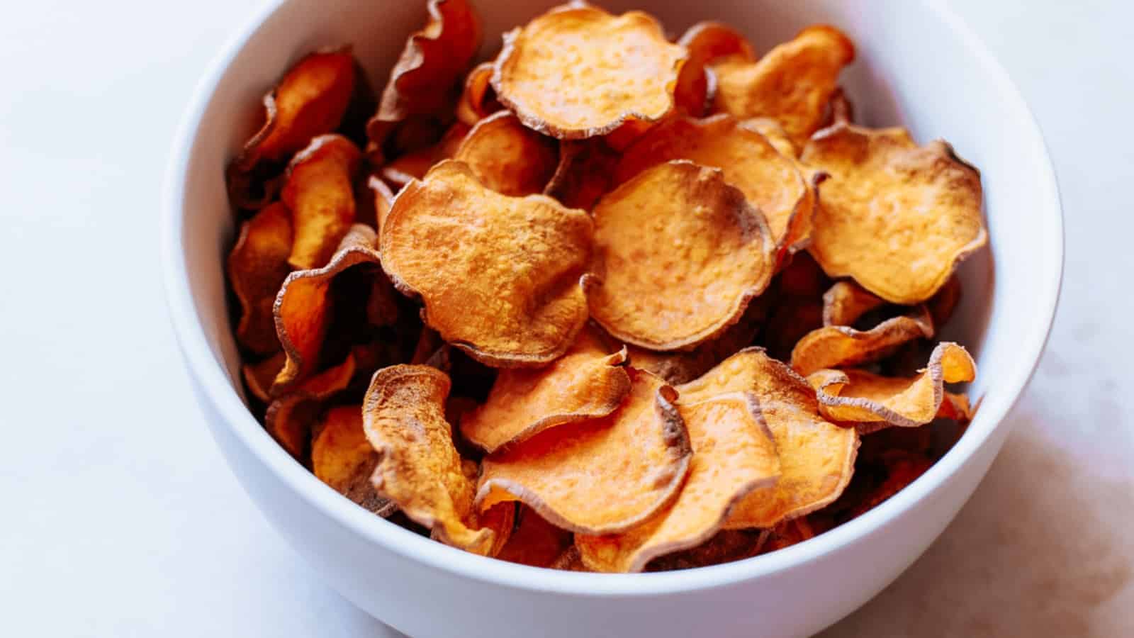 A white bowl filled with crispy sweet potato chips on a light surface. The chips have a golden-brown hue with slightly curled edges.