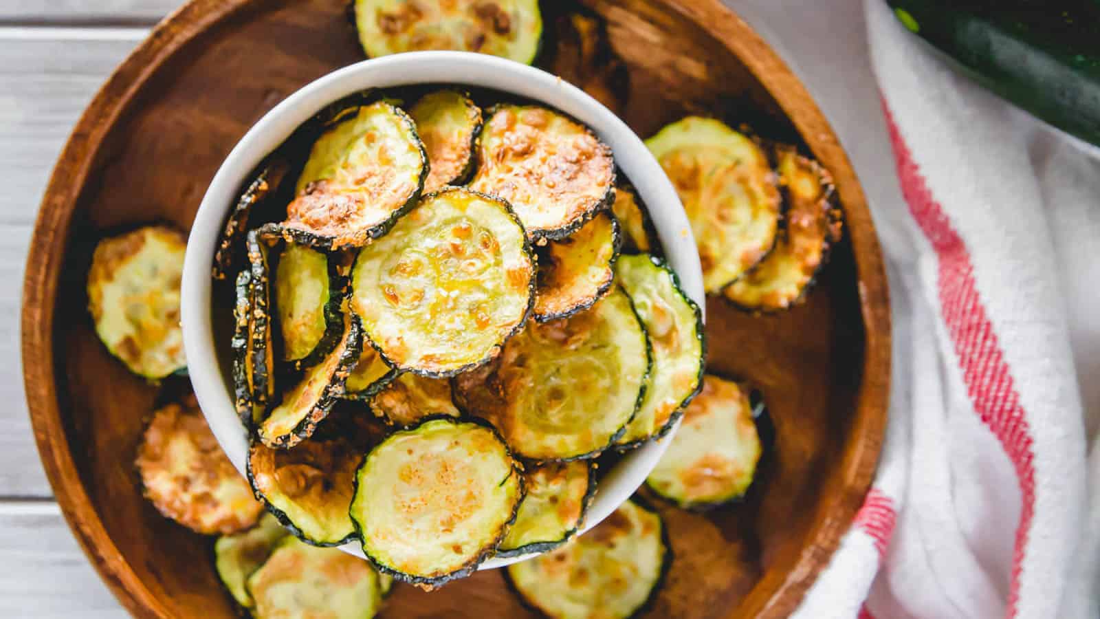 A bowl of crispy zucchini chips is placed on a larger wooden plate filled with more zucchini chips. A white cloth with red stripes is partially visible beside the plate. The chips are thinly sliced and appear to be roasted.