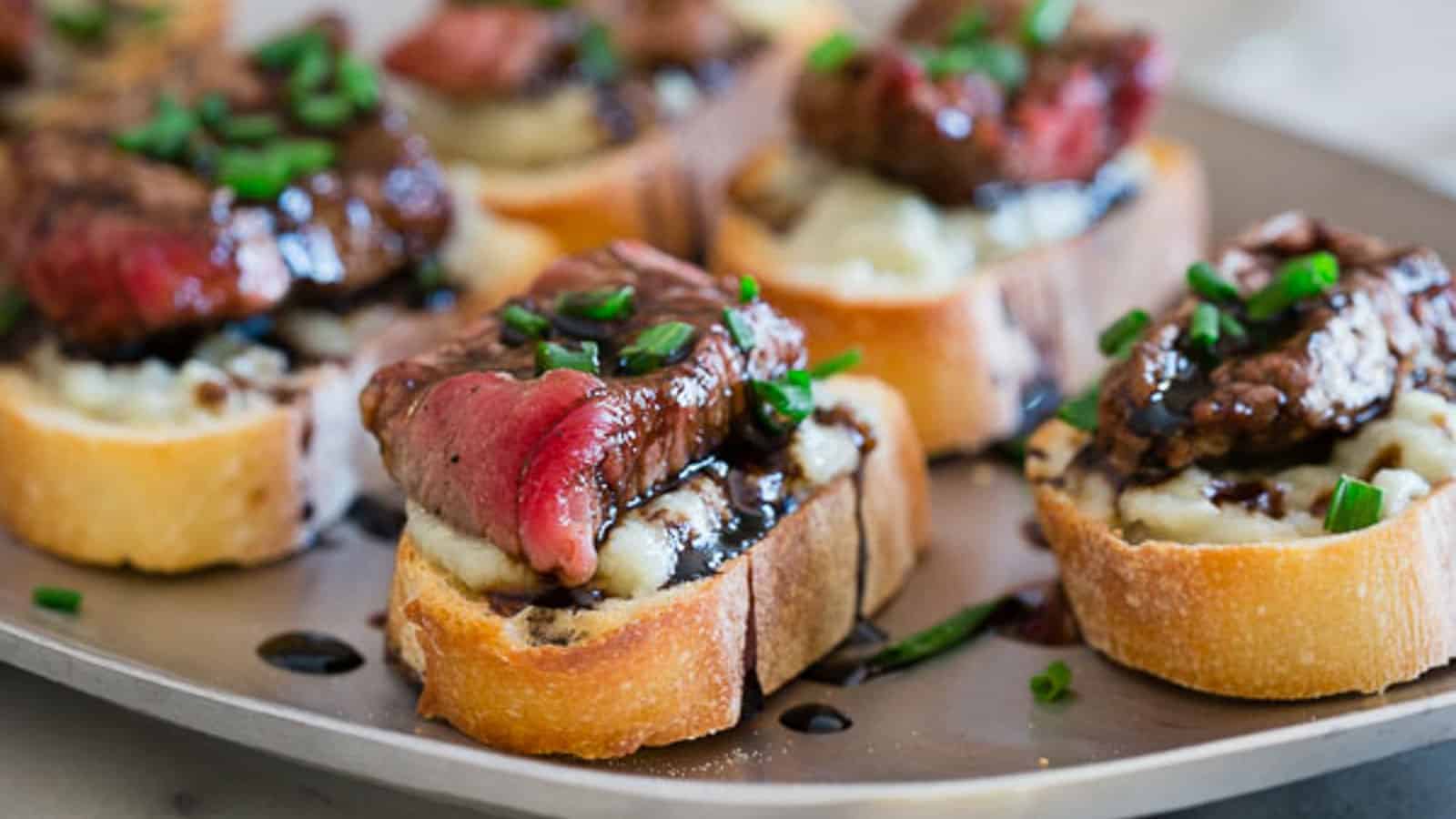 Slices of baguette topped with steak, mashed potatoes, balsamic glaze, and chopped chives are arranged on a plate.