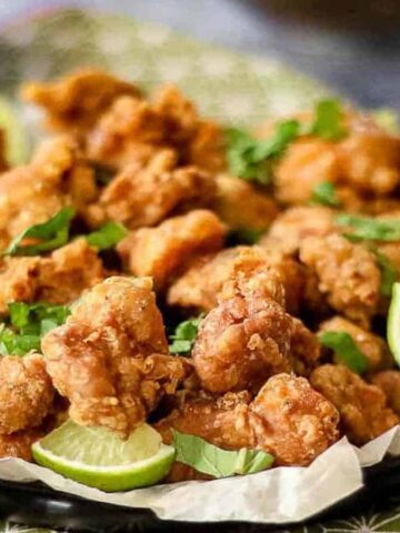 A plate of crispy fried chicken pieces garnished with lime wedges and cilantro. The dish is placed on a green-patterned tablecloth, with small bowls of dipping sauce and other condiments nearby.