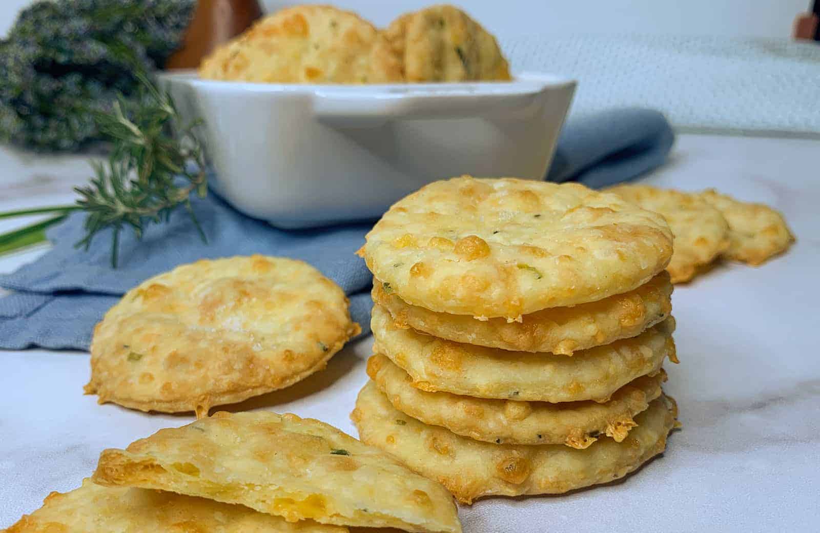 A stack of round, golden-brown cheese crackers on a marble surface. More crackers are scattered around, and a white bowl with additional crackers is in the background. A rosemary sprig and a blue cloth are also visible.