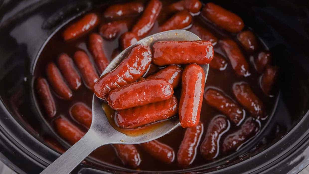 A spoon holds several small sausages coated in a thick sauce, hovering above a pot filled with more sausages in the same sauce.