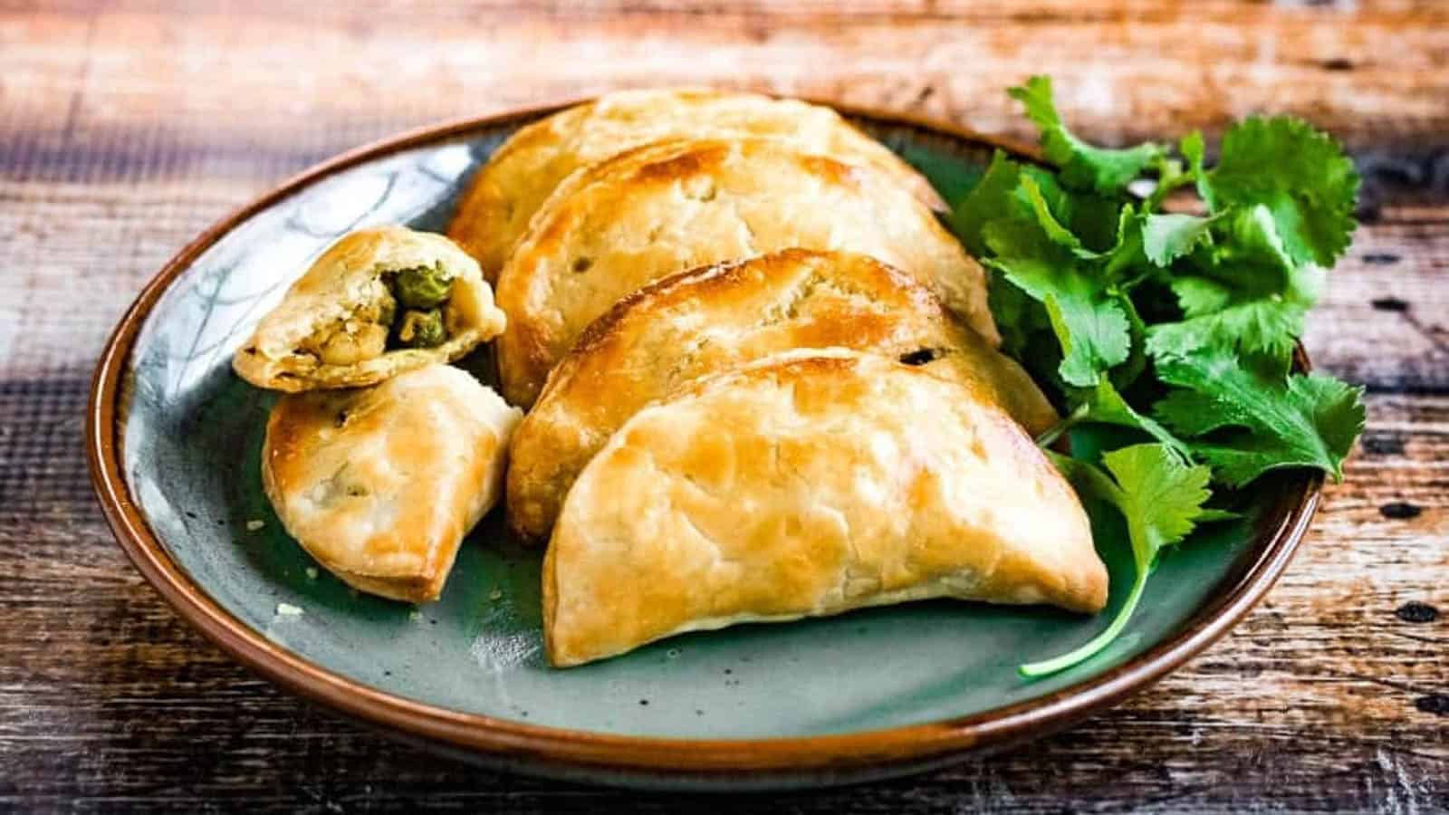 A plate of golden-brown empanadas with one cut open to reveal a vegetable filling. Fresh cilantro is placed beside the empanadas on a rustic wooden table.