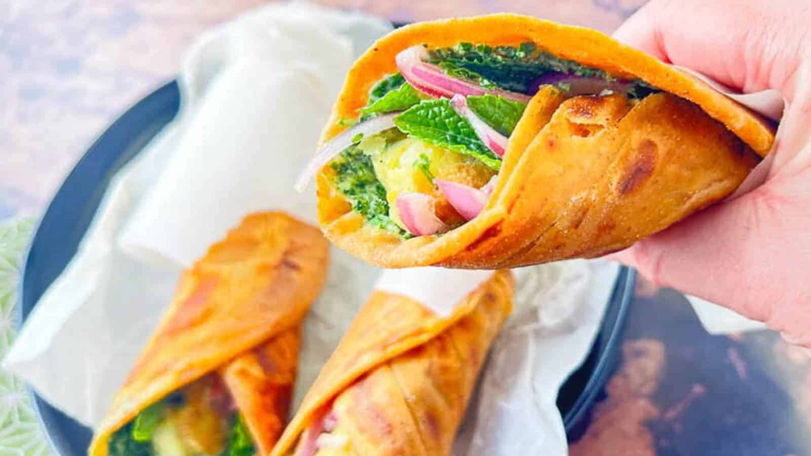 A hand holds a rolled wrap filled with green leafy vegetables, sliced onions, and other ingredients. Two more similarly filled wraps rest on a plate wrapped in paper, set on a light-colored surface.