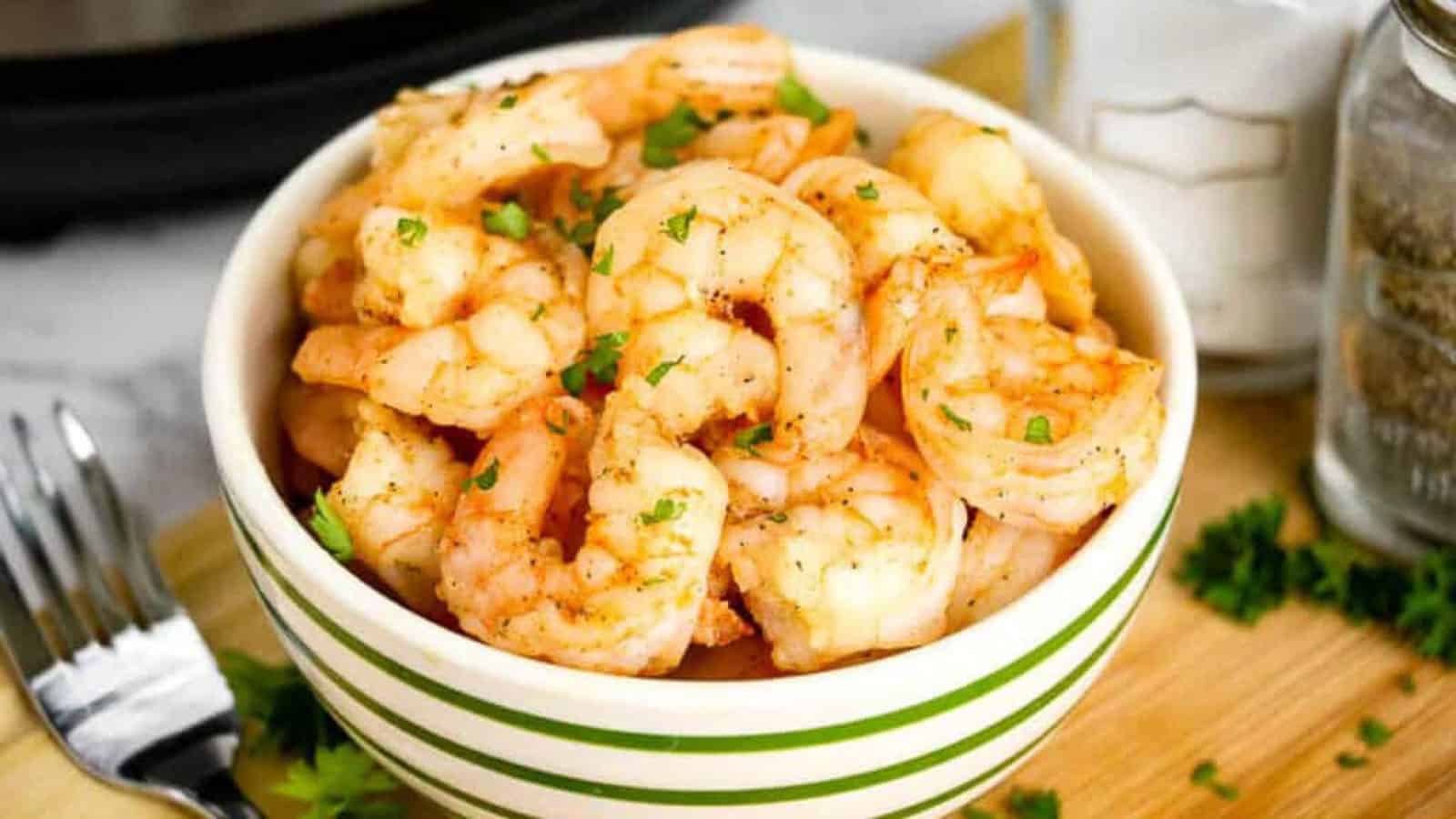 A bowl of cooked shrimp seasoned with herbs is placed on a wooden cutting board. A fork is to the left, and there are two jars in the background, one with salt and the other likely with pepper. Fresh parsley is scattered around.