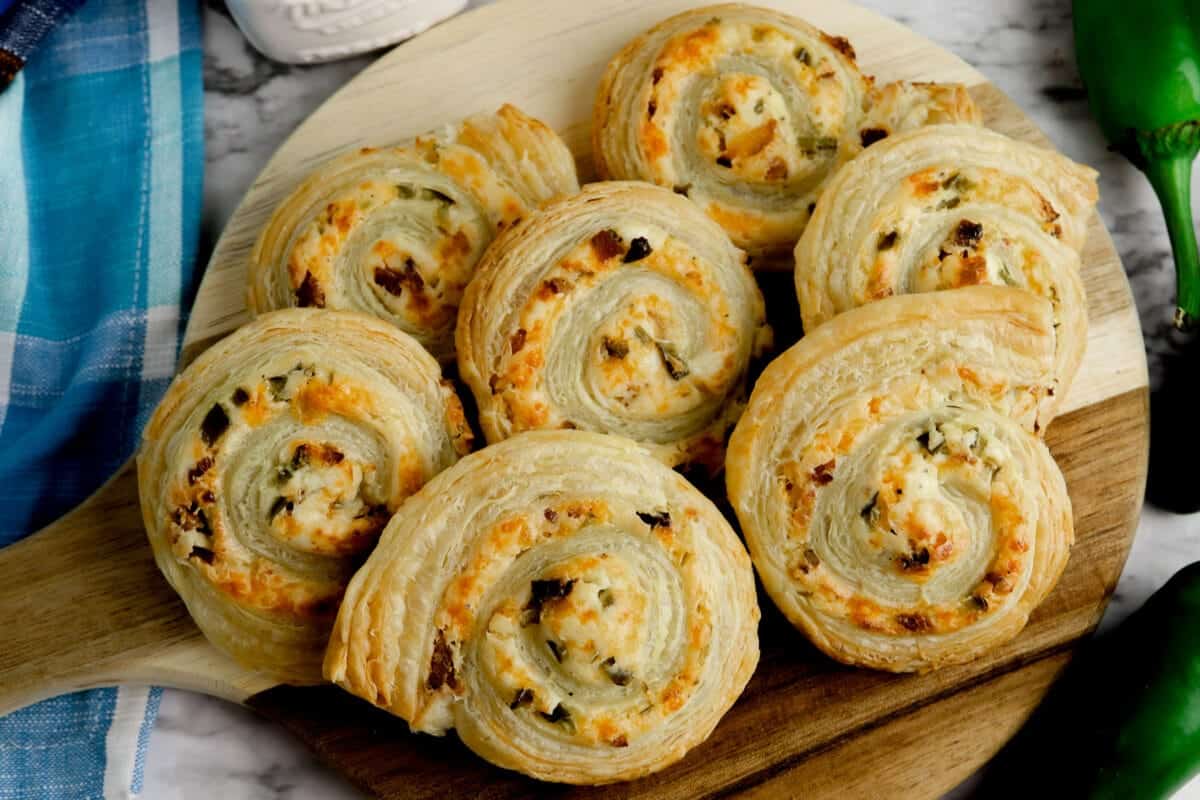 A wooden serving board with several spiral-shaped puff pastries filled with cheese and herbs. The pastries are golden-brown and arranged neatly. A blue cloth and a sliced green chili are partially visible beside the board.